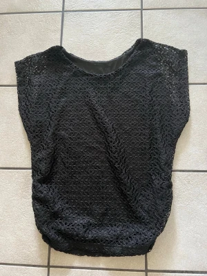 Svart spetsblus med kort ärm storlek L - Snygg svart blus i spets med korta ärmar och rund hals. Blusen har ett vackert hålmönster och är fodrad undertill. Perfekt att styla med jeans eller kjol för en trendig look.