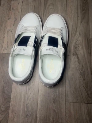 Vita sneakers med svart detalj Valentino - Säljer ett par vita sneakers från Valentino med svart band över vristen och guldfärgad logga på innersulan. Skorna har klassisk låg profil, vita snören och diskreta nitar på hälen. Perfekta för dig som gillar stilrena och exklusiva sneakers.