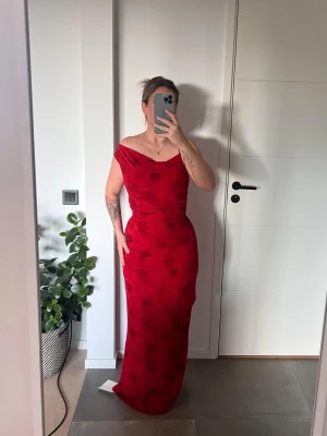 Adoore klänning  - Elegant röd långklänning med offshoulder-design och subtilt blommönster. Klänningen har en figurnära passform och är perfekt för speciella tillfällen. .