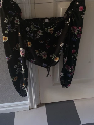 Svart blommig blus med puffärmar - Trendig svart blus med färgglatt blommönster och långa puffärmar. Toppen har smockad överdel och snörning framtill för en snygg passform. Perfekt för dig som vill sticka ut med en romantisk vibe.