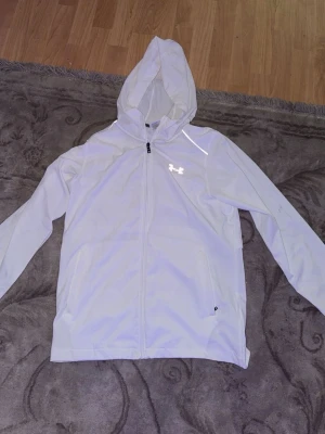 Vit hoodie från Under Armour - Snygg vit hoodie från Under Armour med dragkedja framtill och huva. Hoodien har två sidofickor och en diskret logga på bröstet. Tillverkad i ett lätt och sportigt material som passar perfekt till träning eller chill.