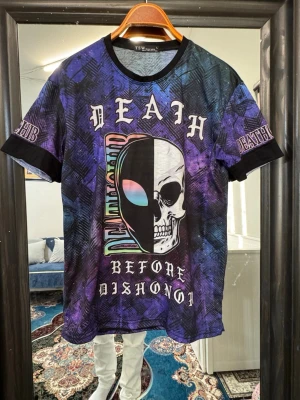 Lila t-shirt med dödskalletryck - Cool t-shirt i lila och blå toner med stort dödskalletryck och texten 'Death Before Dishonor' framtill. T-shirten har korta ärmar, svarta muddar och ett unikt, färgstarkt mönster. Perfekt för dig som gillar streetwear och vill sticka ut.