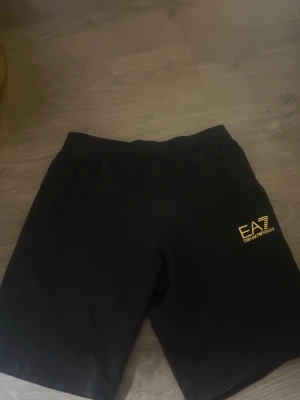 Svarta shorts från Emporio Armani EA7 - Snygga svarta shorts från Emporio Armani EA7 med guld logga på ena benet. Mjuk resår i midjan och två sidofickor. Perfekta för en avslappnad och sportig stil. Tillverkade i ett bekvämt material som passar till sommaren.