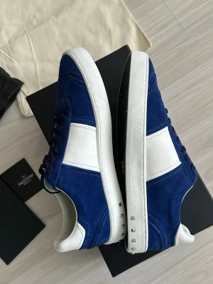Blå och vita sneakers från Valentino - Snygga sneakers i blå mocka och vitt läder från Valentino. Skorna har vita sulor med små silverfärgade nitar på sidan och svarta skosnören. Perfekta för dig som vill ha en stilren men ändå unik look.