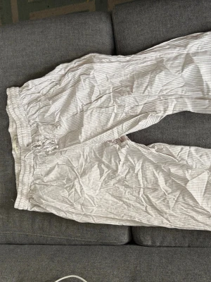 Randiga pyjamasbyxor i bomull - Säljer ett par bekväma pyjamasbyxor med tunna beige och vita ränder. Byxorna har resår i midjan och dragsko för extra komfort. Tillverkade i mjuk bomull, perfekta för slappa dagar hemma eller mysiga kvällar.