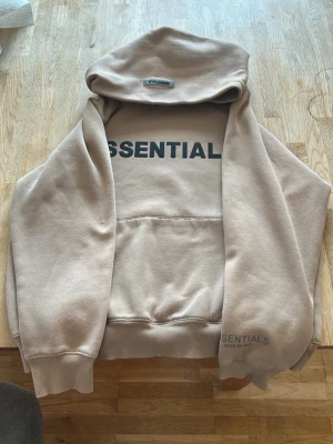 Beige Essentials hoodie med tryck - Säljer en beige hoodie från Essentials med stor svart logga på bröstet och mindre tryck på ärmen. Hoodien har huva och magficka, samt ribbade muddar vid ärmar och nederkant. Tillverkad i mjukt bomullsmaterial som känns riktigt skönt mot huden.