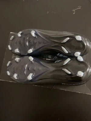Svarta Nike fotbollsskor med blåa detaljer - Snygga svarta fotbollsskor från Nike med högt skaft och blå swoosh på sidan. Skorna har fasta dobbar i vitt och är designade för bra grepp på planen. Lätta och smidiga med snörning för optimal passform. Perfekta för dig som vill ha stilrena och funktionella skor.