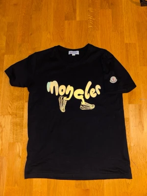 Svart Moncler t-shirt med tryck - Säljer en svart t-shirt från Moncler med coolt tryck framtill där loggan är utformad med sneakers. T-shirten har korta ärmar och klassisk passform. Perfekt för dig som vill ha något unikt och trendigt i garderoben. Passar S och tight på M