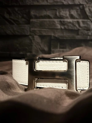 Vändbart bälte med H-spänne - Säljer ett vändbart bälte i läder med en stor, silverfärgad H-formad metallspänne. Ena sidan är vit och den andra svart, vilket gör det enkelt att matcha med olika outfits. Bältet har en stilren och modern design som sticker ut.