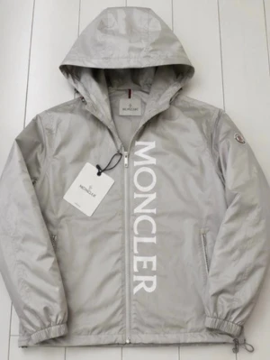 Grå vindjacka från Moncler - Snygg grå vindjacka från Moncler med stor vit logga längs dragkedjan. Jackan har huva, två sidofickor med dragkedja och elastiska muddar vid ärmsluten. Tillverkad i ett glansigt, lätt material som passar perfekt för blåsiga dagar.