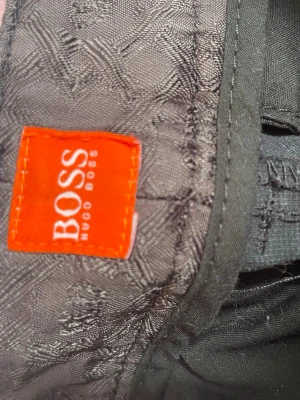 Svarta chinos från Hugo Boss - Snygga svarta chinos från Hugo Boss med klassisk passform. Byxorna har bakficka med knapp och bälteshällor. Tillverkade i ett mjukt och slitstarkt material som ger en clean och stilren look.