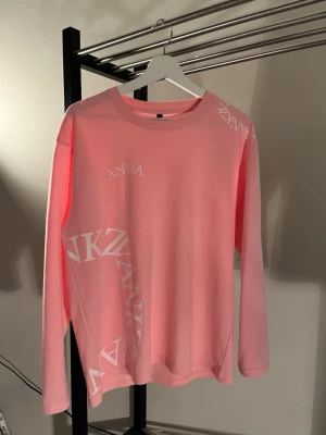Rosa långärmad tröja med tryck - Snygg rosa långärmad tröja med vita grafiska tryck på bröstet, axeln och sidan. Tröjan har en avslappnad passform och rund halsringning. Perfekt för dig som gillar streetwear och vill sticka ut med färg.