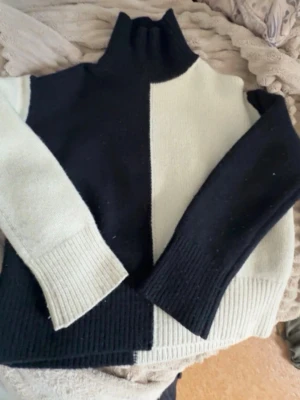  Trendig Svart och vit stickad polotröja - Säljer en trendig stickad polotröja i svart och vitt blockmönster. Tröjan har hög krage och långa ärmar med breda ribbade muddar. Perfekt för dig som gillar en stilren och modern look.