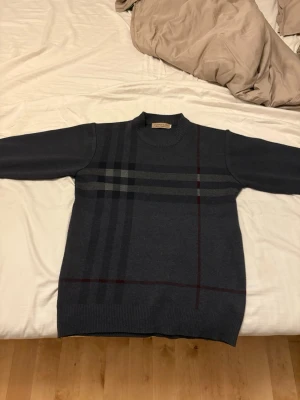 Mörkgrå stickad tröja  - Snygg mörkgrå stickad tröja med klassiskt rutmönster i svart, grått och vinrött. Tröjan har rund halsringning och ribbade muddar. Perfekt för dig som gillar stilrena och tidlösa plagg.