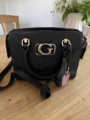 Svart handväska Guess med axelrem - Snygg svart handväska från Guess med både handtag och justerbar axelrem. Väskan har silverfärgade detaljer, stor G-logga framtill och dragkedjefack på baksidan. Tillverkad i konstläder och har en stilren, boxig form. Perfekt för dig som gillar trendiga accessoarer.