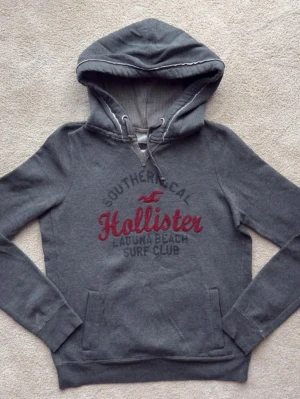 Grå hoodie från Hollister med tryck - Mysig grå hoodie från Hollister med broderat rött och svart tryck på bröstet där det står 'Hollister Laguna Beach Surf Club'. Hoodien har huva med dragsko, känguruficka och ribbade muddar. Tillverkad i bomull och polyesterblandning.
