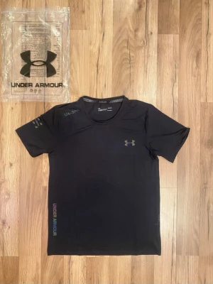 Svart tränings-tshirt från Under Armour - Svart t-shirt från Under Armour med korta ärmar och rund hals. Tillverkad i ett lätt och stretchigt syntetmaterial, perfekt för träning. Diskret logga på bröstet och tryck på ärmen samt sidan. Snygg och sportig design.