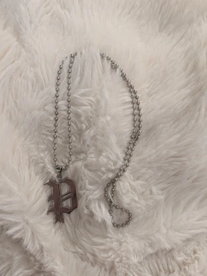 Kedjehalsband med bokstavshänge - Snyggt halsband i silverfärgad metall med ett stort bokstavshänge i gotisk stil. Kedjan består av små runda pärlor och hänget har en unik design som sticker ut. Perfekt accessoar för dig som vill ha något personligt och edgy.