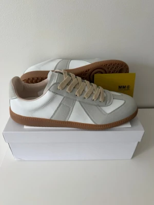 Vita och grå sneakers med gummisula - Snygga sneakers i vitt och ljusgrått med beige snören och klassisk gummisula. Ovandelen är i en mix av läder och mocka för en stilren look. Perfekta för dig som gillar minimalistisk och tidlös design.