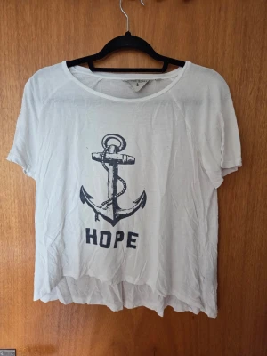 Vit t-shirt med ankare  - Vit t-shirt med ett stort svart ankare och texten HOPE på framsidan. T-shirten har rund halsringning och korta ärmar. Tillverkad i mjuk bomull som känns skön mot huden. Perfekt för en avslappnad och stilren look.