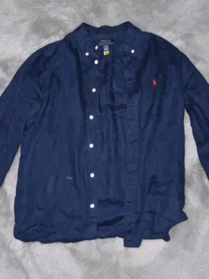 Marinblå skjorta från Polo Ralph Lauren - Klassisk marinblå skjorta från Polo Ralph Lauren med vita knappar och röd broderad logga på bröstet. Skjortan har lång ärm och är tillverkad i ett mjukt bomullstyg. Perfekt för en stilren och avslappnad look.