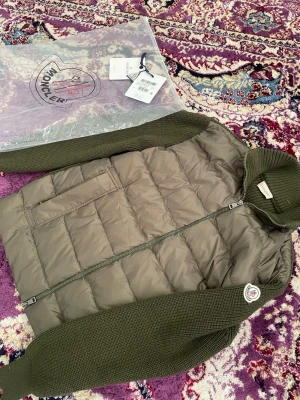 Moncler cardigan  - Snygg olivgrön dunväst från Moncler med stickade ärmar och hög krage. Västen har quiltad front, dragkedja och två fickor. Moncler-logga på ärmen och detaljer i matchande grönt. Perfekt för lager-på-lager och streetstyle. Helt ny och har även nfc Scan.