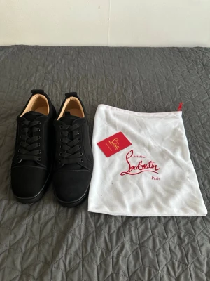  Christian Louboutin - Snygga svarta sneakers från Christian Louboutin med klassisk röd sula och svarta skosnören. Skorna har en stilren design i mocka och läderdetaljer, samt ikonisk branding på sulan. Perfekta för dig som vill ha en exklusiv och trendig look.