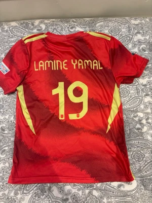 Spansk landslagströja Lamine Yamal - Säljer en röd och gul matchtröja från Adidas för Spaniens landslag med Lamine Yamal och nummer 19 på ryggen. Tröjan har korta ärmar, officiella tryck och HEAT.RDY-material för extra komfort. Perfekt för fotbollsfans som vill stötta Spanien.