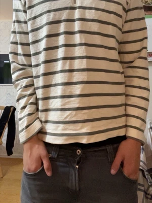 Randig tröja med halv dragkedja - Snygg långärmad tröja med vita och mörkgrå ränder samt halv dragkedja framtill. Tröjan har en hög krage i kontrasterande mörkgrå färg och är tillverkad i ett mjukt bomullsliknande material. Perfekt för en avslappnad och stilren look.