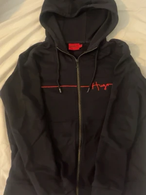 Svart hoodie från Hugo Boss - Säljer en svart hoodie från Hugo Boss med dragkedja framtill och röd Hugo-logga broderad på bröstet. Passar definitivt S, pris kan diskuteras