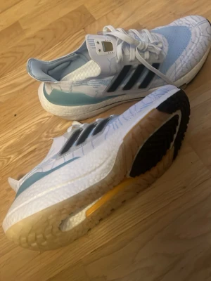 Vita Adidas sneakers ultra boost  - Snygga vita Adidas sneakers med blå och svarta detaljer. Ovandelen är i mesh och syntet, med klassiska tre svarta ränder på sidan. Sulan är tjock och har en gul detalj undertill. Skorna har vita snören och en bekväm passform. Argentina collab 
