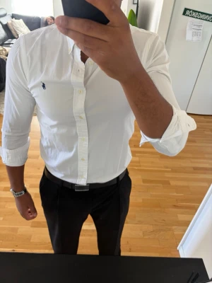 Vit skjorta från Ralph Lauren - Klassisk vit skjorta från Ralph Lauren med knappslå och broderad logga på bröstet. Skjortan har lång ärm, smal passform och är tillverkad i bomull. Perfekt till både jeans och kostymbyxor.