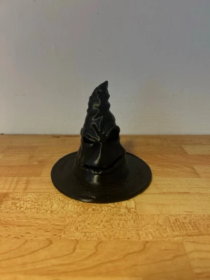 Svart trollkarlshatt cosplay - Cool svart trollkarlshatt i plast med detaljerat ansikte och spetsig form. Perfekt för maskerad, cosplay eller temafest. Hatten har en bred brätte och ett hål undertill för enkel användning. Lägg ett fale ljus i hålet så börjar den lysa från munnen och ögonen. 