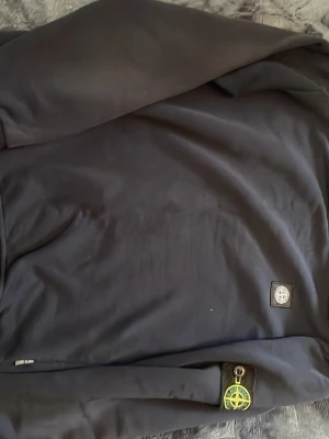 Mörkblå sweatshirt från Stone Island - Säljer en mörkblå sweatshirt från Stone Island med klassisk rund hals och ikonisk patch på vänster ärm. Tröjan har en stilren design med diskret logga på bröstet och är tillverkad i mjukt bomullsmaterial. Perfekt för en avslappnad och trendig look. Storlek S pris kan diskuteras