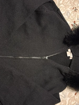 Svart jacka med fuskpäls och dragkedja - Svart jacka med huva och mjuk fuskpäls runt huvkanten. Jackan har dragkedja framtill och är långärmad. Perfekt för kyligare dagar och har en stilren look som passar till det mesta.