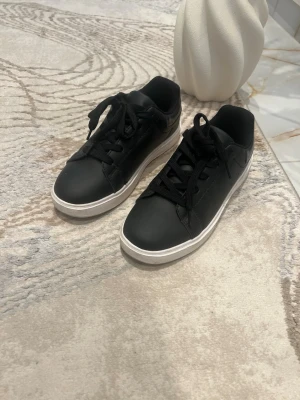 Svarta sneakers med vit sula - Stilrena svarta sneakers med klassisk låg profil och vit sula. Skorna har svarta skosnören och ovandel i slätt material som ger en clean look. Perfekta för dig som gillar enkel och tidlös stil.