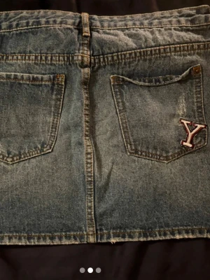 Jeanskjol med patches och broderier - Cool jeanskjol i blå denim med flera broderade patches framtill och en Y-bokstav på bakfickan. Kjolen har klassisk femficksdesign, bälteshällor och dragkedja. Perfekt för dig som gillar en personlig och trendig stil.
