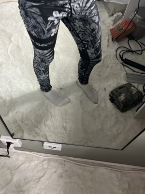Stronger - Snygga svarta leggings med vitt och grått blommönster och textdetalj i midjan. Tight passform och stretchigt material som sitter skönt. Perfekta för dig som vill ha något bekvämt men ändå trendigt.