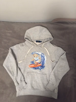 Polo Bear Ralph Lauren - Säljer en grå hoodie från Polo Bear by Ralph Lauren med en stor färgglad print av en surfande björn på bröstet. Hoodien har dragsko i huvan och ribbade muddar. Perfekt för dig som gillar streetwear och vill ha något unikt och bekvämt.