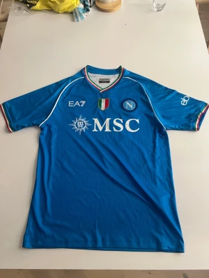 Napoli 22/23 hemma Di Lorenzo #22 - Snygg blå Napoli matchtröja från EA7 med Di Lorenzo och nummer 22 på ryggen. Tröjan har vita detaljer, klubbmärke och sponsorer på bröstet samt italienska flaggans färger i krage och ärmslut. Perfekt för fotbollsfans som vill sticka ut på läktaren.