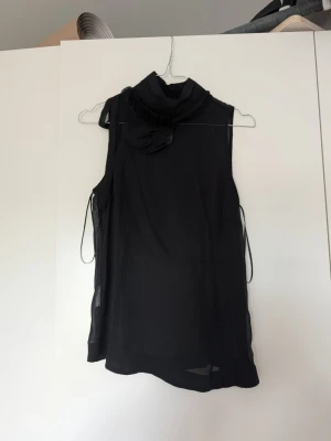Svart ärmlös blus med rosett - Säljer en svart, ärmlös blus i ett tunt och lätt material. Blusen har hög krage med en dekorativ rosett och volangdetalj vid halsen. Perfekt för dig som gillar en elegant och stilren look.