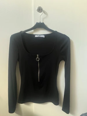 Svart ribbad topp med dragkedja - Snygg svart långärmad topp från NA-KD med ribbad struktur och en cool silverfärgad dragkedja framtill. Toppen har en rundad urringning. Det är en xxs tröja men passar både som S och Xs för den är stretchig