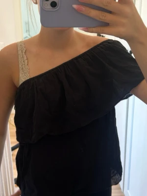 Svart oneshoulder topp med volang - Säljer en svart oneshoulder topp med volangdetalj längs överkanten. Toppen är ärmlös och har en luftig passform