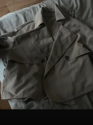 Kort beige trenchcoat med bälte - Snygg kort beige trenchcoat med bred krage och dubbelknäppning. Jackan har ett matchande bälte i midjan och dekorativa knappar framtill. Tillverkad i ett slätt tyg som ger en klassisk look. Perfekt för dig som vill ha en stilren och trendig jacka.