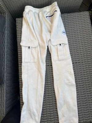 Beiga cargobyxor från Under Armour - Snygga beiga cargobyxor från Under Armour med stora fickor på sidorna och broderad logga på ena benet. Tillverkade i mjukt, stretchigt material som ger en avslappnad och sportig look. Perfekta för dig som gillar streetwear och funktionella detaljer.