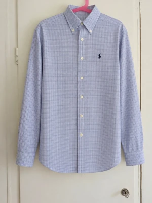 💙 Polo Ralph Lauren blå rutig skjorta – storlek 12 - Klassisk och stilren skjorta från Polo Ralph Lauren i ljusblått med diskret rutigt mönster. Button-down-krage, långa ärmar och den ikoniska mörkblå broderade loggan på bröstet. Storlek: 12 år Färg: Ljusblå/vit Skick: Fint begagnat skick men den har några fläckar! Väldigt lättstylad och passar både till vardags och finare tillfällen. Snygg för en klassisk preppy/Stockholm-stil. Skriv gärna om du vill ha mått!