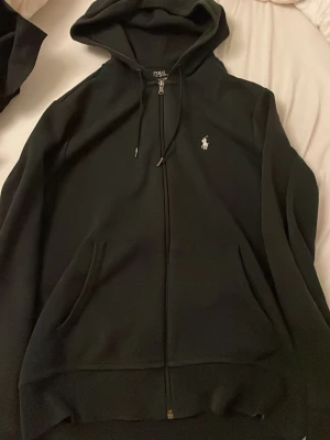 Svart hoodie från Polo Ralph Lauren - Klassisk svart hoodie från Polo Ralph Lauren med dragkedja framtill, två fickor och vit broderad logga på bröstet. Huva med dragsko och mjukt material som känns skönt mot huden. Perfekt för en avslappnad och stilren look.