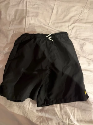 Svarta badbyxor från Lyle & Scott - Säljer ett par svarta badbyxor från Lyle & Scott Junior med elastisk midja och vit dragsko. Klassisk modell med gul logga på benet och meshfoder på insidan. Perfekta för stranden eller poolen.