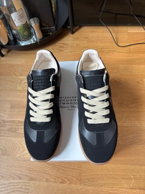 Maison Margiela svarta sneakers - Maison Margiela sneakers i svart läder och mocka med vita skosnören och gummisula. Klassisk low-top modell med stilren design och diskret logotyp på plösen. Perfekta för dig som gillar minimalistisk och trendig stil.