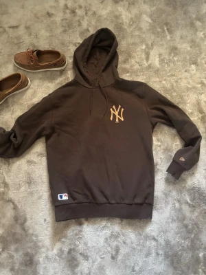 Brun New York Yankees hoodie - Snygg brun hoodie med New York Yankees-logga i beige på bröstet. Klassisk huva med dragsko och ribbade muddar vid ärmar och nederkant. MLB-märke nertill och diskret broderad logga på ärmen. Perfekt för dig som gillar sportig streetwear.